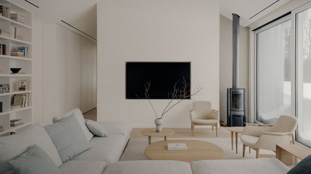 Bright, minimalist living room with a large pale sectional sofa, built-in white shelving and concealed cabinetry, a wall-mounted television, nested light-wood coffee tables, upholstered lounge chairs, a freestanding wood-burning stove, and full-height glazed doors or windows with sheer curtains. The room has a calm neutral palette and clean-lined detailing.