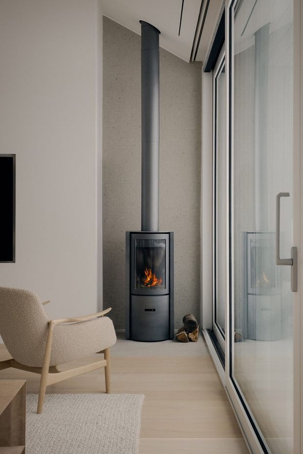 A refined, minimalist interior corner featuring a freestanding black wood-burning stove with a tall flue set against a small beige mosaic-tile feature wall. Full-height glazed doors or windows line the right side, while a light upholstered lounge chair, pale timber flooring, and a neutral textured rug create a calm seating area. A partial wall-mounted screen is visible at left.