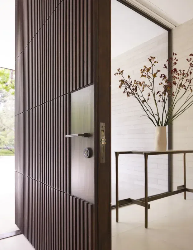 A close-up view of a refined residential entryway featuring a tall dark timber pivot door with vertically fluted detailing, a frosted glass inset, and contemporary hardware. Beyond the door is a bright foyer with white horizontal wall cladding, a slim dark console table, and a tall neutral vase with branches. The composition emphasises the architectural entrance and the contrast between dark timber and light finishes.
