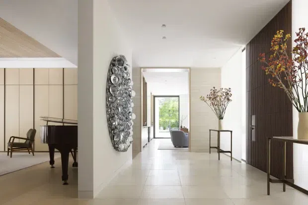 Bright, refined residential entry hall with a long tiled floor and sightline into a living area. The space features white walls, a pale timber-lined ceiling section, recessed lighting, a large sculptural wall artwork, a slim console table with floral arrangement, and dark vertical timber screening with integrated joinery on the right. A piano and lounge seating are visible in adjoining rooms.