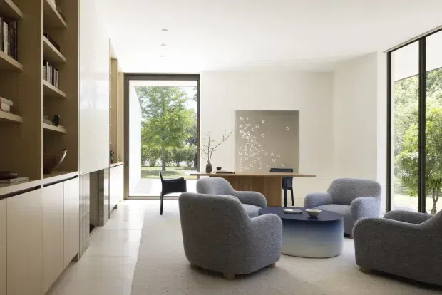 Bright, minimalist living room with floor-to-ceiling glazing and views of greenery. The room features curved grey upholstered lounge chairs around a low rounded coffee table, a long timber dining or console table, pale flooring, and full-height built-in shelving and cabinetry along the left wall. A restrained neutral palette and clean architectural detailing create an airy contemporary interior.