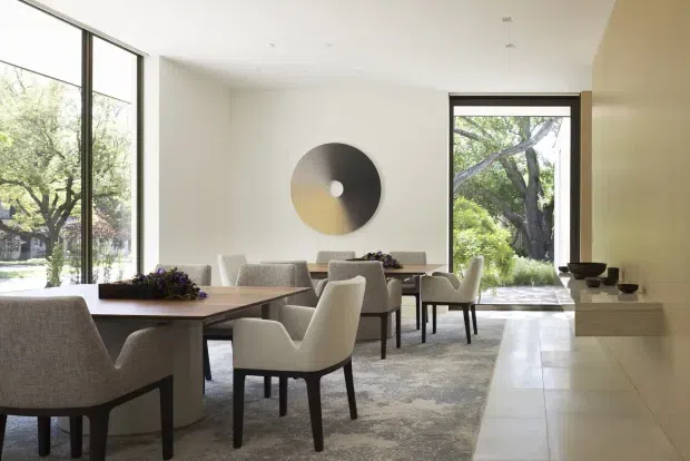 Bright, refined open-plan dining area with a long timber dining table, upholstered dining chairs, large floor-to-ceiling windows, and a glazed garden-facing doorway. A pale tiled circulation zone runs alongside a softly patterned area rug, while a minimalist circular wall artwork and simple built-in display ledge add understated detail.
