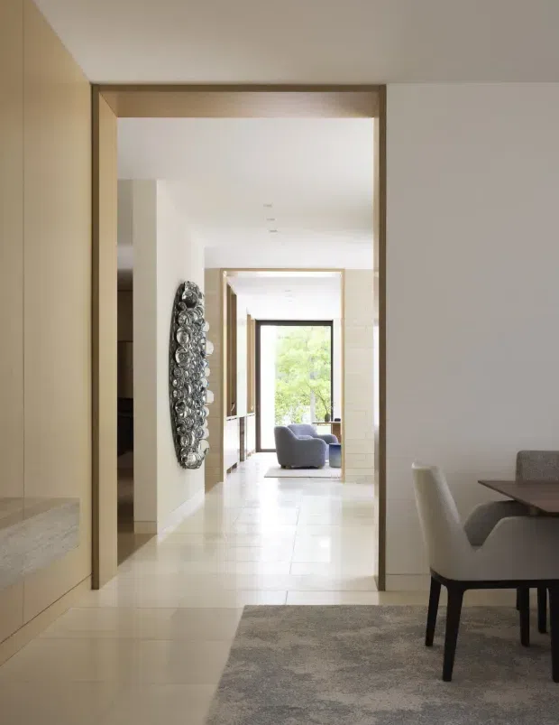 A bright, refined interior view looking through a sequence of wide, timber-trimmed openings into a sitting area with an armchair and large garden-facing window. The foreground includes a dining area with a dark tabletop, upholstered chairs, built-in pale cabinetry, light tiled flooring, and a textured grey rug. A sculptural metallic wall artwork provides a focal point along the hallway. The space has clean lines, abundant natural light, and a calm neutral palette.