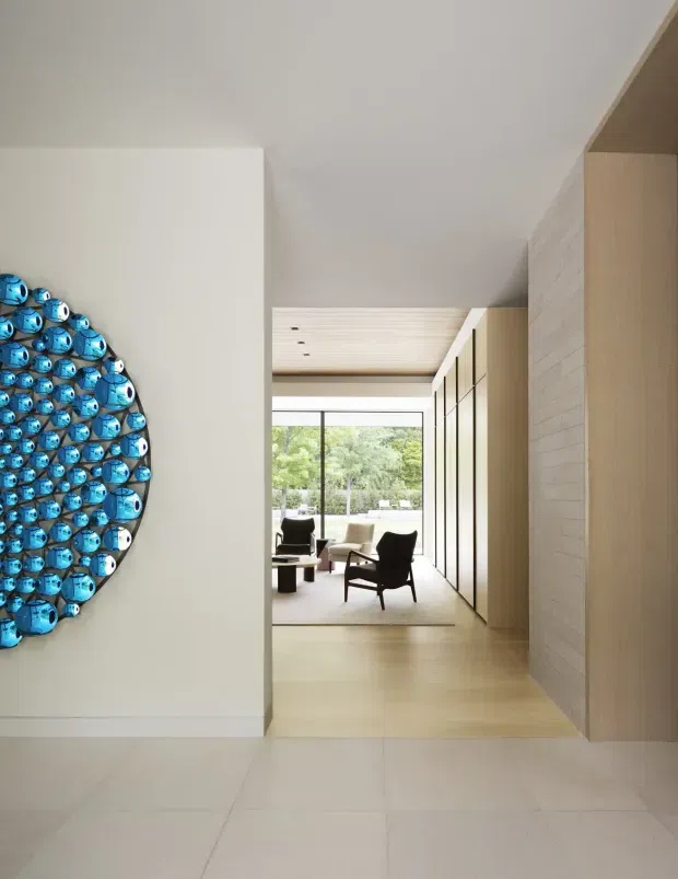 A refined, minimalist residential interior viewed from a pale tiled entry or circulation space toward a bright living room. The composition features warm light-wood flooring and ceiling lining, full-height light timber built-in cabinetry, a pale textured masonry or stone feature wall, and large glazed doors opening to greenery. A striking circular blue sculptural wall installation provides the main visual accent, while two dark lounge chairs and a low coffee table sit on a neutral rug in the living area.