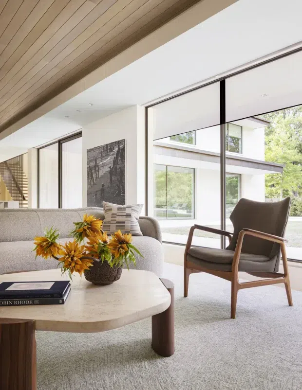 A bright, refined modern living room with expansive floor-to-ceiling glazing, a neutral sofa, sculptural rounded coffee table, and a mid-century-inspired timber-framed lounge chair. A timber-lined ceiling adds warmth, while the windows overlook a landscaped courtyard and a modern building exterior. The room is softly furnished with a textured rug, cushions, artwork, books, and a floral arrangement.