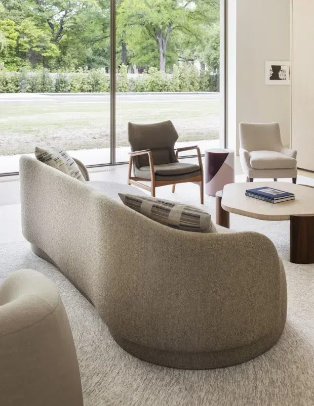 A bright, refined living room with a large curved upholstered sofa in a soft beige boucle-like finish, patterned lumbar cushions, a dark wood and cream round coffee table, sculptural side table, and two mid-century-inspired lounge chairs. Full-height glazing opens to a leafy garden, while pale walls and carpet create a calm, neutral interior.