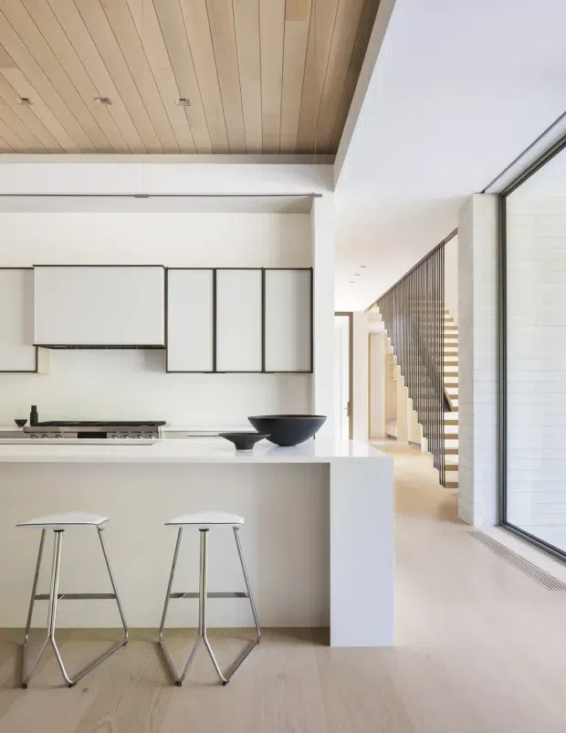 Bright minimalist kitchen with a white island, integrated white cabinetry outlined by dark frames, gas cooktop, and two streamlined metal-and-white stools. Pale timber boards continue across the floor and ceiling, while a large glazed opening brings in abundant daylight. A stairway with slender dark vertical balustrading is visible beyond the kitchen.