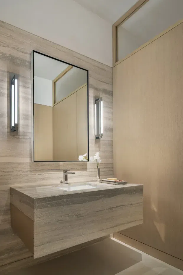 A refined contemporary bathroom vanity with a floating stone-clad basin unit, integrated white basin, chrome tapware, large frameless mirror, and slim vertical wall lights. The walls and vanity feature horizontally veined beige-grey stone, while full-height light timber cabinetry and an upper interior window add warmth and visual depth. Soft concealed lighting beneath the vanity enhances the clean, luxurious composition.