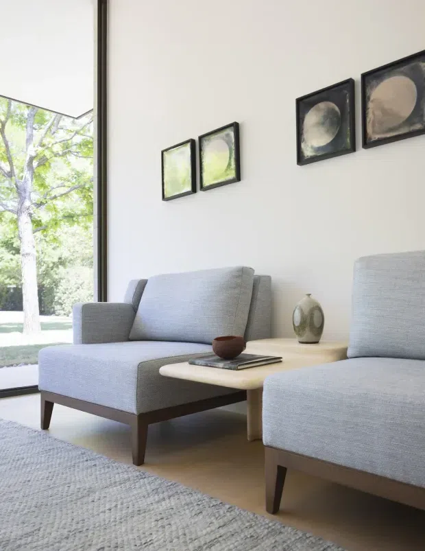 Bright, calm living room with two upholstered grey armchairs arranged around a light wood or pale stone coffee table. A large floor-to-ceiling window brings in views of trees and natural light. Minimal framed abstract artwork hangs on the white wall, with a textured neutral rug and understated ceramic accessories completing the refined contemporary setting.