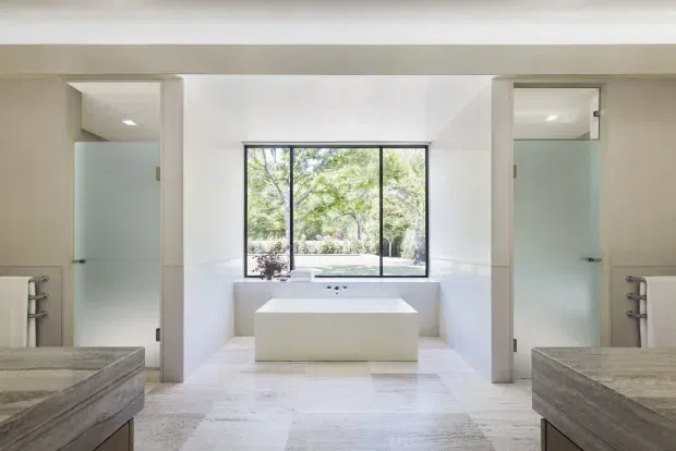 Bright, symmetrical contemporary bathroom with a freestanding rectangular bathtub positioned beneath a large black-framed window overlooking greenery. Two frosted-glass shower enclosures flank the tub, with matching wall-mounted towel rails and stone-look vanity edges visible in the foreground. The room features predominantly white finishes, pale stone surfaces, clean geometric lines, and abundant natural light.