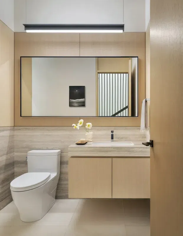 A compact, refined contemporary bathroom with a wall-mounted timber vanity, integrated white basin, large black-framed mirror, toilet, stone-look wall finish, and pale tiled floor. Warm timber joinery and neutral stone surfaces are balanced by a linear pendant light and a small floral arrangement. A stair or screened opening is reflected in the mirror.