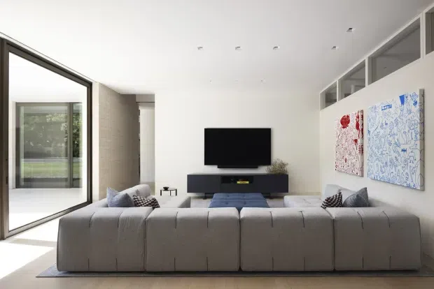 A bright, minimalist living room viewed from behind a large modular sectional sofa. The room features a neutral grey sofa, low dark media console, wall-mounted television, pale flooring, white walls and ceiling, expansive black-framed glazing on the left, and high-level interior windows along the right wall. Abstract artwork adds restrained colour.
