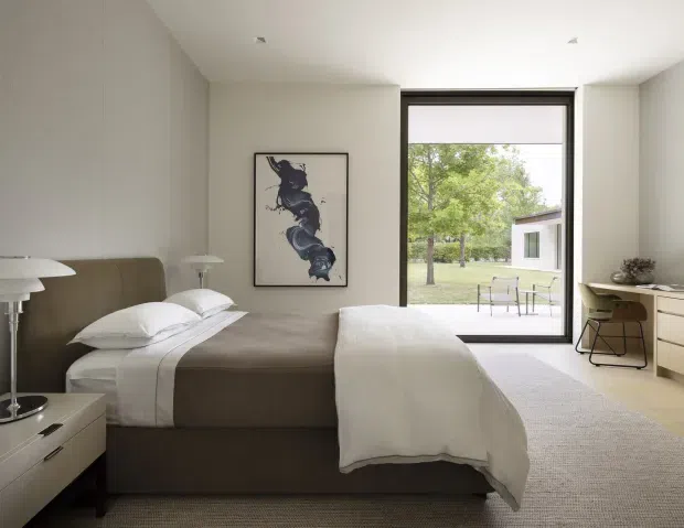 A serene, minimalist bedroom with a low upholstered taupe bed, crisp white bedding, matching bedside tables and lamps, and a pale textured carpet. A large black-framed glazed door opens to a landscaped outdoor area and a separate modern pavilion. Neutral walls, restrained furnishings, and a monochromatic artwork create a calm, refined atmosphere.