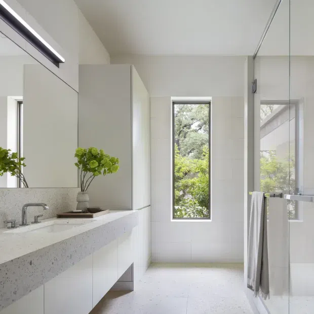 Bright contemporary bathroom with a long floating white vanity, integrated pale terrazzo benchtop, wall-to-wall mirror, frameless glass shower enclosure, and a narrow vertical window overlooking greenery. The room has clean white tiled walls, pale terrazzo flooring, minimalist chrome tapware, and soft natural light.