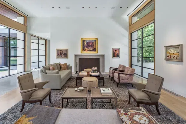A bright, refined living room arranged around a central fireplace, with a large sectional sofa, upholstered armchairs, ottomans, layered coffee tables, and a patterned area rug. Tall black-framed windows and glazed doors bring in abundant natural light, while warm timber-framed clerestory windows and pale walls create a balanced contemporary-traditional setting. Artwork is displayed around the fireplace and on the surrounding walls.