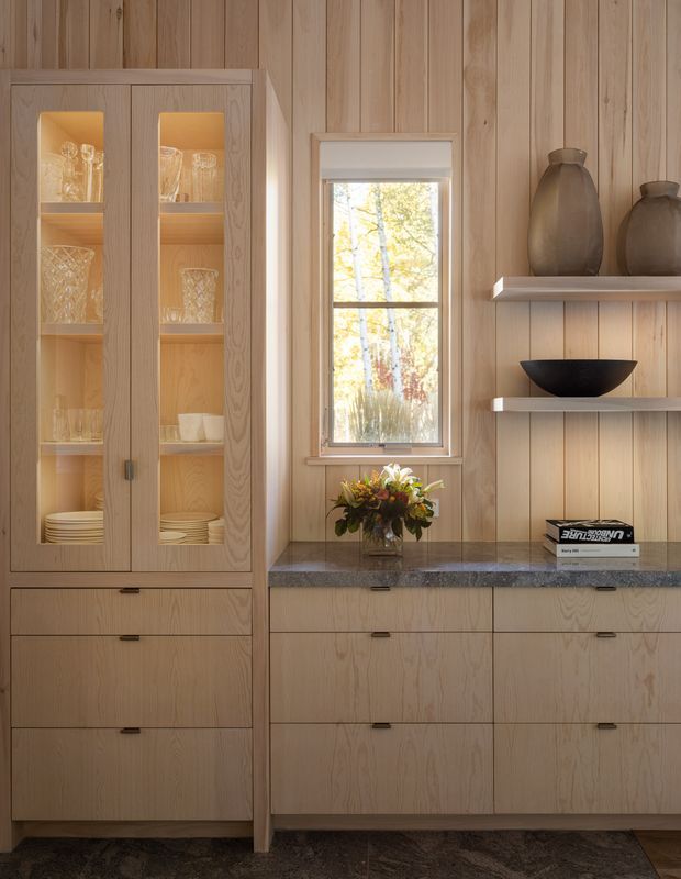 Warm, refined built-in cabinetry and display storage in a wood-panelled room. Light timber joinery with glass-fronted doors, drawers, open display shelves, a dark stone benchtop, and decorative ceramics and glassware surrounds a central window.