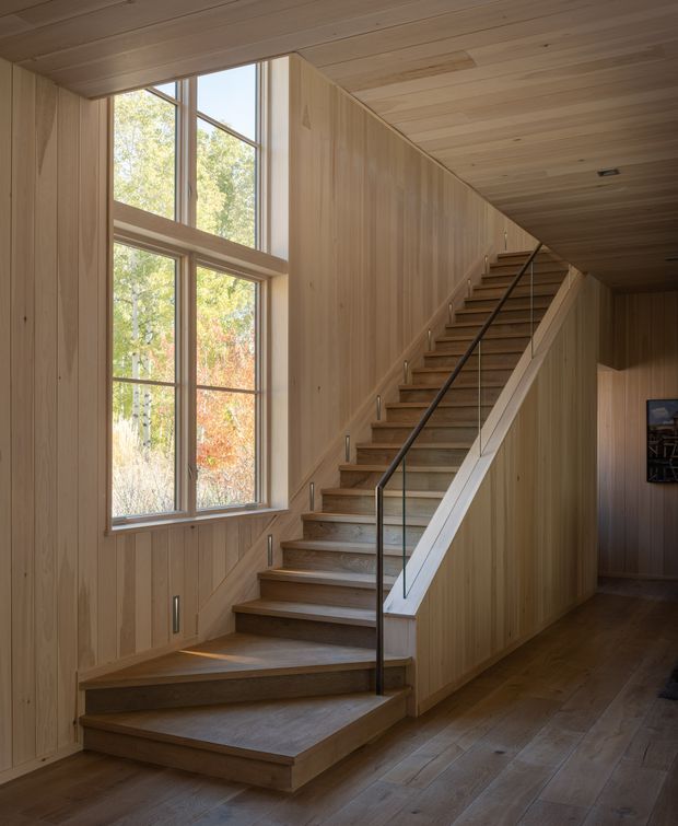A warm, wood-lined stairway rises beside tall multi-pane windows overlooking trees. The stairs and surrounding walls, ceiling, and flooring are finished in natural timber, with a slim dark metal handrail and understated recessed lighting. The space has a simple, highly integrated contemporary cabin-like character.