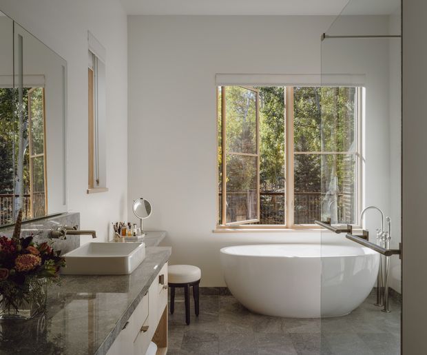 A bright, refined bathroom with a freestanding white bathtub positioned beneath large timber-framed windows, a broad stone vanity with a vessel basin, wall mirror, and polished floor tiles. The room overlooks leafy greenery and uses a calm neutral palette with natural wood accents.
