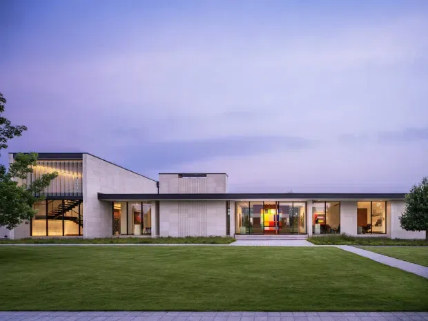 A low, elongated modern building viewed from across a broad manicured lawn at dusk. The facade combines pale masonry or concrete-like wall panels, extensive floor-to-ceiling glazing, a slim dark flat roof, and a taller glazed volume on the left with visible internal stairs and vertical screening. A brightly coloured abstract feature is visible through the central entrance glazing, with paved paths leading toward the building.
