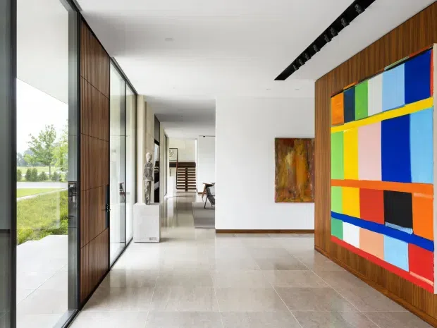 A bright, elongated contemporary interior hallway or entry gallery with full-height glazing on one side, a large timber pivot door, polished pale stone flooring, white walls, and warm vertical timber wall panelling. A colourful geometric artwork dominates the right wall, while a linear black ceiling light and additional artwork add visual interest. The space opens toward a dining or living area in the distance, with landscaped grounds visible through the glazing.