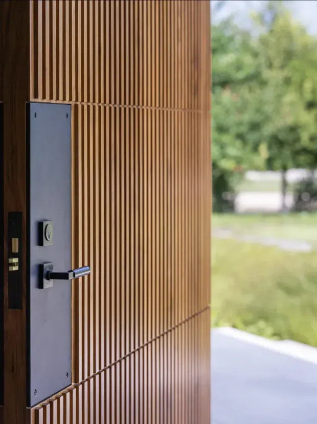 Close-up view of a contemporary exterior entry door with a warm timber slatted finish, a tall brushed-metal lock plate, and a sleek horizontal metal lever handle. The door is open to a blurred green outdoor setting and pale paved entry area.