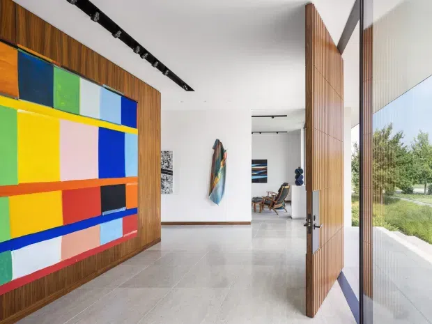 A bright contemporary entry hall with a large colourful geometric artwork mounted on a timber-paneled wall. The space has pale stone or porcelain flooring, white walls, black track lighting, a tall timber pivot door, and full-height glazing opening toward landscaped grounds. A living area with lounge seating is visible beyond the hall.