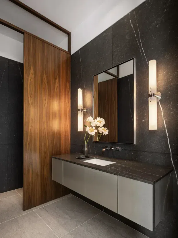 A refined contemporary bathroom featuring a floating double vanity with grey cabinetry, a dark veined stone benchtop and matching full-height wall finish, a rectangular mirror, vertical wall sconces, and a tall warm timber door or partition. Large-format grey floor tiles and dark metal fixtures complete the high-contrast scheme.