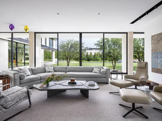 A bright, spacious contemporary living room with a large grey sectional sofa, sculptural stone or concrete-look coffee table, patterned lounge chairs, and a matching upholstered armchair with ottoman. Floor-to-ceiling black-framed glazing opens views to a landscaped courtyard and neighbouring modern architecture. The room has a white ceiling, pale neutral carpet, exposed light-toned structural columns, and a large textured wall artwork or feature surface.