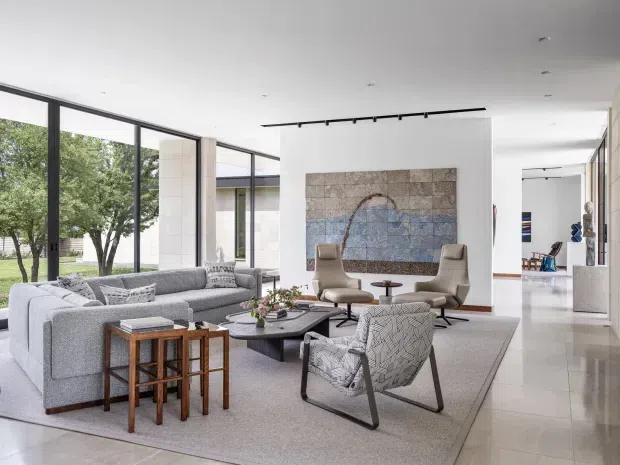 Bright, spacious contemporary living room with floor-to-ceiling black-framed glazing, a large sectional sofa, sculptural lounge chairs, a low coffee table, and a large abstract wall artwork. The room has a pale neutral palette, polished light flooring, recessed ceiling lights, track lighting, and views to a landscaped garden. An open circulation area and additional interior spaces are visible beyond the main lounge.
