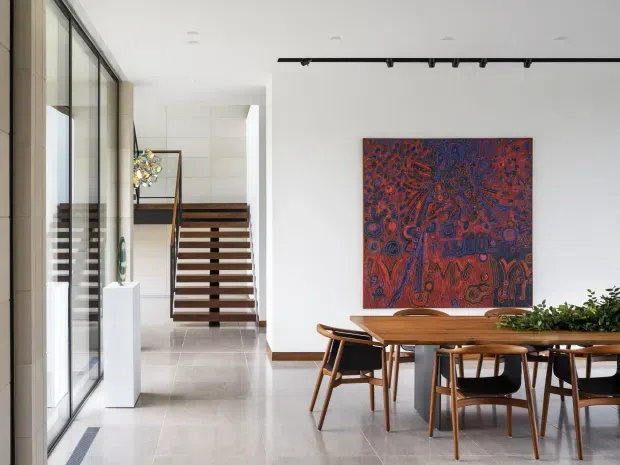 A bright, refined open-plan dining area adjoining a stairway. The space features a long timber dining table with matching timber-and-dark upholstered chairs, a large colourful artwork on a white wall, a linear black track light, pale large-format tiled flooring, full-height glazed sliding doors on the left, and a floating timber staircase with a dark metal or timber handrail. A planter runs along the rear of the dining table, while a sculptural object is displayed near the stairs.
