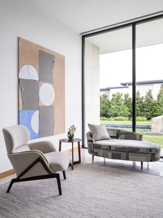 A bright, refined contemporary living room with floor-to-ceiling black-framed glazing overlooking a landscaped garden and swimming pool. The room features a sculptural upholstered lounge chair, a curved modular chaise, a small dark wood side table, a large abstract wall artwork, and a pale textured carpet. White walls and large-format light flooring create an airy, minimalist setting.