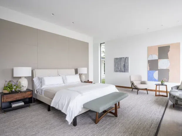 Bright, spacious contemporary bedroom with a low-profile upholstered bed positioned against a full-height taupe upholstered or paneled feature wall. White bedding, bedside tables with ceramic lamps, a large pale grey area rug, and a muted sage upholstered bench create a calm, refined scheme. A seating corner, abstract artwork, and tall narrow glazing are visible in the background.