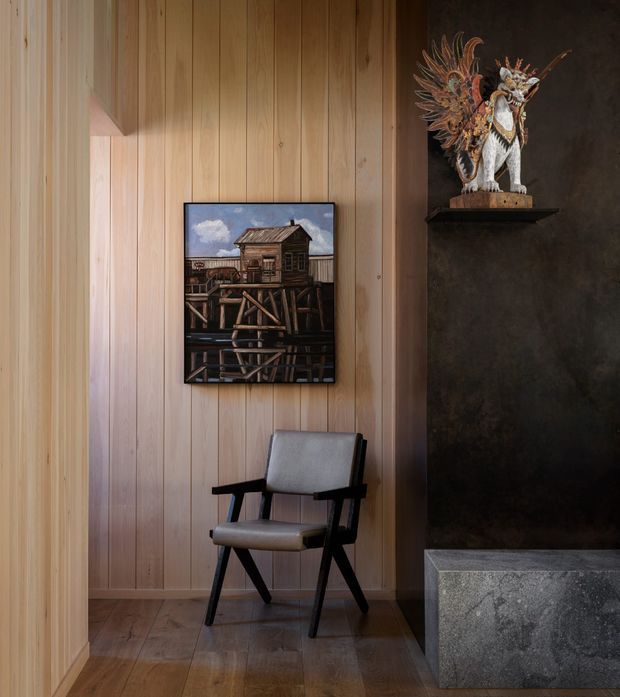 A warm, moody interior nook with vertical light timber wall panelling, timber flooring, and a dark textured feature wall. A mid-century-style upholstered armchair sits beneath a framed artwork, beside a sculptural decorative object displayed on a shelf above a substantial grey stone hearth or plinth.