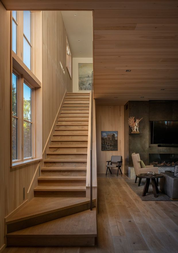 A warm, wood-lined interior showing a straight staircase beside tall timber-framed windows, opening onto a living room with a fireplace, television, lounge seating and timber flooring. The walls, ceiling, staircase and window surrounds are extensively finished in natural wood, creating a cohesive contemporary cabin-like atmosphere.
