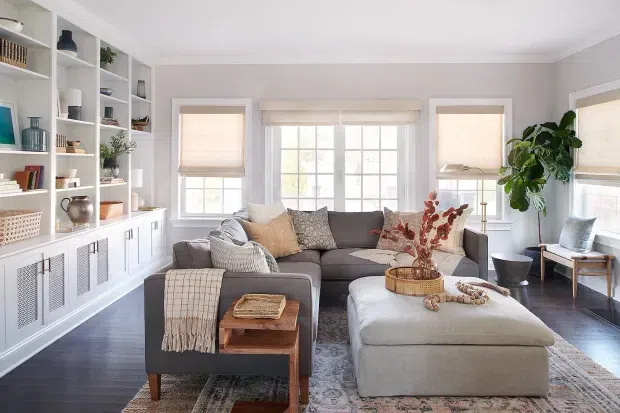 A bright, carefully styled living room with a large grey sectional sofa and matching ottoman, layered neutral cushions, a patterned area rug, and warm wood accents. Built-in white shelving and lower cabinetry line the left wall, while multiple windows with light Roman shades provide abundant natural light. A large indoor plant and understated decorative accessories add an inviting, refined feel.
