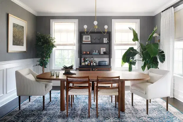 A refined dining room with a long mid-century-style timber dining table surrounded by upholstered armchairs and wooden dining chairs. The room features dark grey vertical wall panelling above white wainscoting, white trim and ceiling, large windows with sheer curtains, a patterned area rug, a dark built-in or freestanding shelving unit, indoor plants, and a brass multi-arm pendant light.