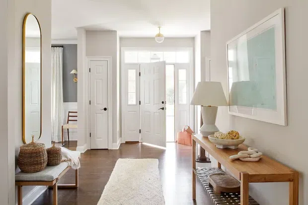 Bright, welcoming residential entry hall with white paneled doors, a glazed front door, warm timber flooring, a long light-coloured runner, and a narrow timber console styled with a lamp and decorative objects. A tall arched mirror, woven baskets, bench seating, artwork, and soft curtains add a relaxed, refined look. The space opens toward a dining area and is illuminated by abundant natural light.