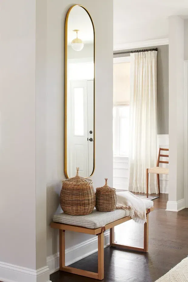 Bright, refined entryway featuring a tall arched mirror with a thin brass-gold frame, a timber bench with a light upholstered cushion, woven lidded baskets, white painted walls and trim, and dark timber flooring. A curtained adjoining room and a wooden chair are visible beyond the entry.