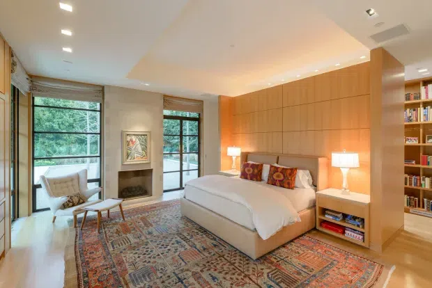 Warm, refined bedroom with a low upholstered bed set against a full-height timber-panelled feature wall. Floor-to-ceiling windows and glazed doors bring in abundant natural light and overlook greenery, while a patterned area rug, decorative cushions, bedside lamps, built-in shelving and a compact fireplace create a comfortable, library-like retreat.