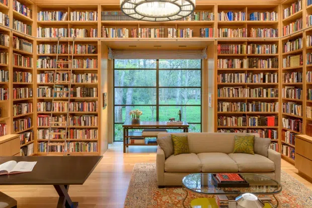 Warm, richly detailed home library or study with floor-to-ceiling built-in timber bookshelves covering multiple walls, a large central window overlooking greenery, and a comfortable seating area with a beige sofa, patterned rug, glass coffee table, and dark wood desk. A decorative circular chandelier hangs from the ceiling, creating an inviting and refined atmosphere.
