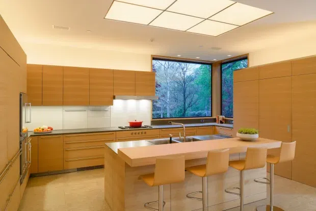 A warm, contemporary kitchen with extensive light-to-mid timber cabinetry, stainless steel appliances, pale stone benchtops, and a large central island with integrated seating. Floor-to-ceiling corner glazing overlooks trees, while a large recessed rectangular ceiling light provides soft illumination. The space appears spacious, highly organised, and designed for both cooking and casual dining.