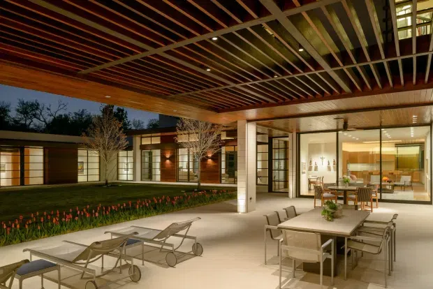 A sophisticated covered outdoor terrace and courtyard at dusk, featuring a large dining table with chairs, lounge seating, integrated lighting, and a dramatic exposed timber-and-steel slatted roof. The terrace opens onto a landscaped courtyard and connects to a brightly glazed contemporary interior with kitchen and dining areas visible inside.