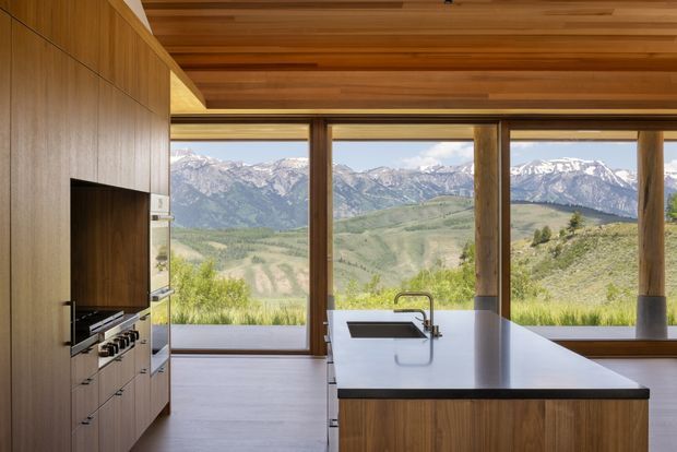 A contemporary kitchen with extensive floor-to-ceiling glazing framing a dramatic mountain landscape. The room features warm timber wall cabinetry, a timber-lined ceiling, integrated appliances, and a large island with a pale benchtop and undermount sink. The strong connection to the landscape and natural materials create a refined, calm interior.