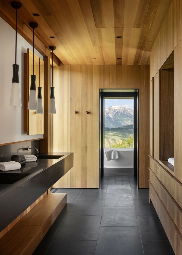 A refined contemporary bathroom clad extensively in warm natural timber, with a floating dark stone vanity, integrated basin, black tapware, pendant lights, full-height timber cabinetry, and dark large-format floor tiles. A freestanding or partially visible bath is seen through the doorway, with a dramatic mountain landscape beyond the glazing.