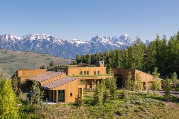 A low-rise contemporary timber-clad building complex set within a natural, lightly landscaped site. The interconnected wings have simple pitched and flat roof forms, warm wood façades, large glazed openings, and sheltered outdoor circulation. A dramatic alpine mountain range is visible in the background.
