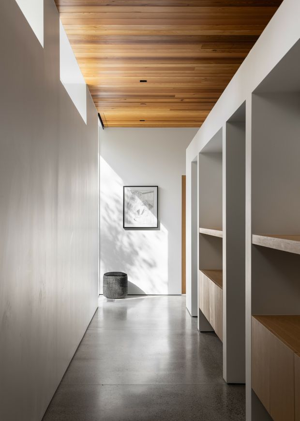 A minimalist residential hallway with a long polished concrete floor, white walls, integrated open shelving and timber cabinetry along one side, and a warm timber-lined ceiling. Natural light creates soft shadows on the walls, with a framed artwork and small upholstered stool at the far end.