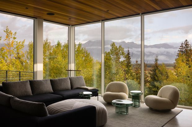 A contemporary living room with a large dark sectional sofa, rounded lounge chairs, glass coffee tables, and a sculptural ribbed ottoman. Full-height corner glazing provides expansive views of a wooded landscape and distant mountains. The room features a warm timber-lined ceiling, slim dark window frames, and a neutral textured rug.