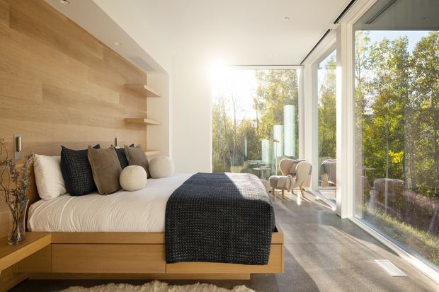 Bright, serene contemporary bedroom with a built-in timber platform bed and integrated bedside storage, a full-height timber feature wall with floating shelves, and expansive floor-to-ceiling glazing overlooking leafy trees. Neutral bedding, textured dark throw, sculptural cushions, and two armchairs create a warm, refined retreat.