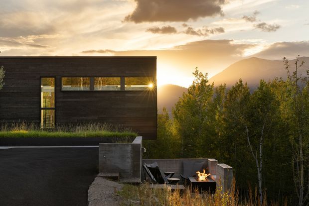 A modern dark-clad house set on a wooded, mountainous site at sunset. The image focuses on a simple rectangular building volume with narrow horizontal glazing and a tall vertical window, overlooking a recessed outdoor terrace with concrete walls, lounge chairs, and a small fire pit. Dense trees and a mountain backdrop reinforce the secluded alpine setting.
