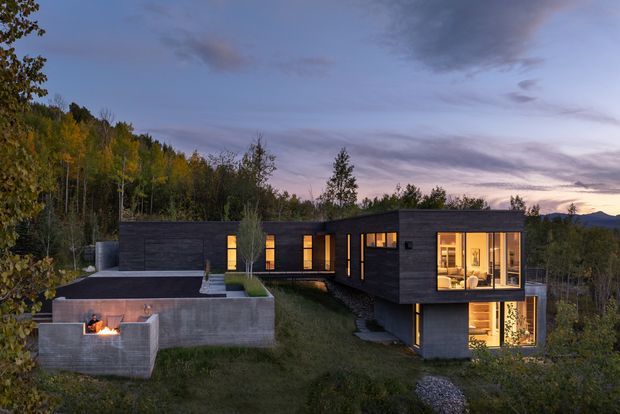 A contemporary single-storey, low-profile home set within a wooded, mountainous landscape at dusk. The building is composed of dark horizontal-clad rectangular volumes with extensive warm-lit glazing. A concrete terrace and outdoor fire feature sit to the left, while a projecting glazed room and lower-level illuminated space create a layered composition across the sloping site.